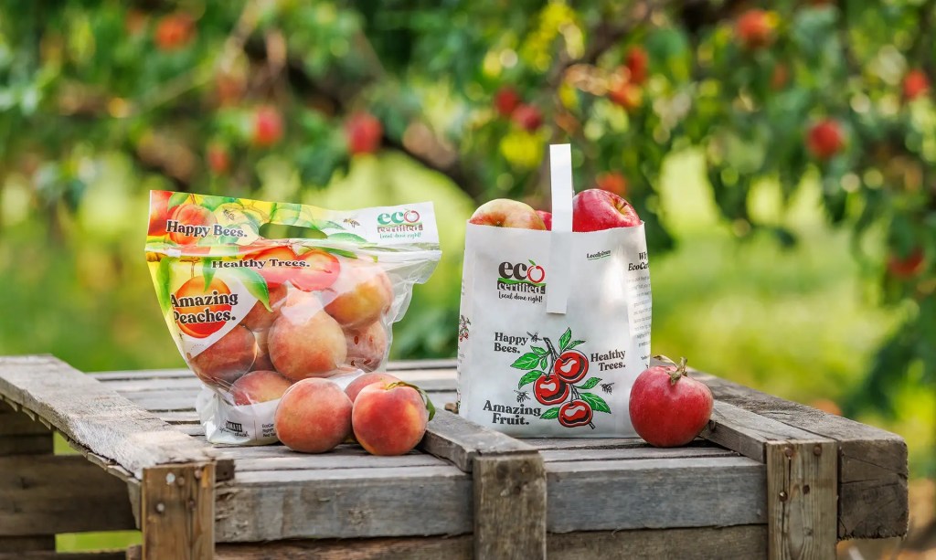 A package of EcoCertified peaches and an EcoCertified Apple tote sit side-by-side on a wooden apple crate, with an orchard in the background